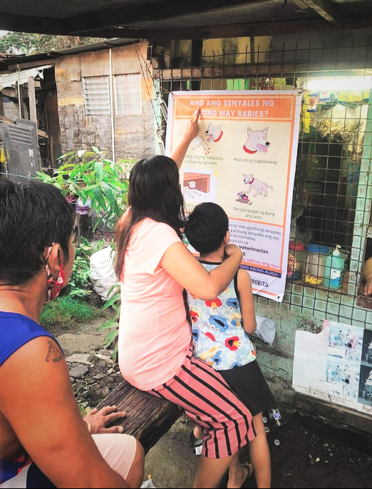 A mother and child read a GARC rabies awareness poster as they wait in line at a local shop. 